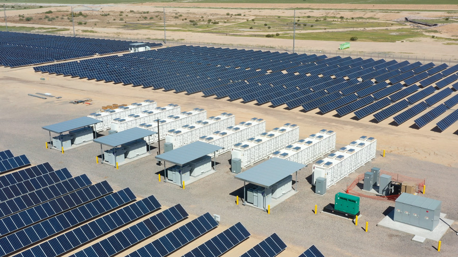 Rows of solar panels beside grid-scale battery storage containers at a desert energy facility.